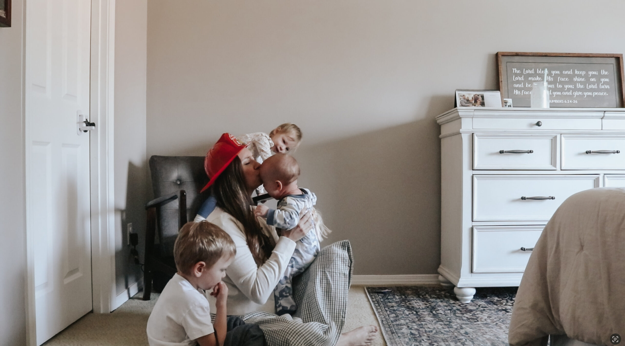 Homemaker is wearing a toy fire hat while kissing her child on the cheek. Two other children are playing next to her.